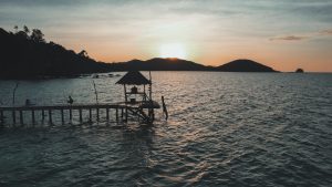 Scenic sunset view at a wooden pier in Ko Mak, Thailand, overlooking the sea and hills.